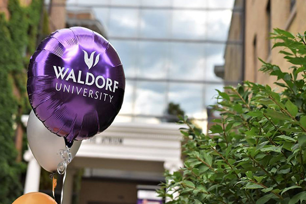Purple mylar balloon with Waldorf University logo on it floating outside with the Atrium in the distance