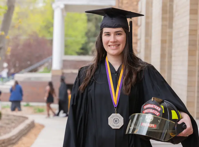 Graduating female in cap & gown holding a fireman's helmet